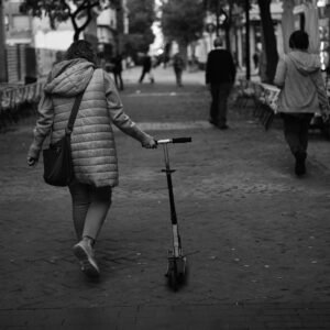 Mujer de espaldas caminando despacio por una calle peatonal mientras arrastra un patinete, rodeada de terrazas vacías y viandantes.