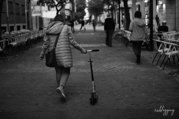 Mujer de espaldas caminando despacio por una calle peatonal mientras arrastra un patinete, rodeada de terrazas vacías y viandantes.