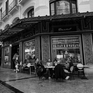 Grupo de personas sentadas en la terraza de una cafetería gourmet en una esquina señorial, bajo una marquesina con reloj y fachada decorada