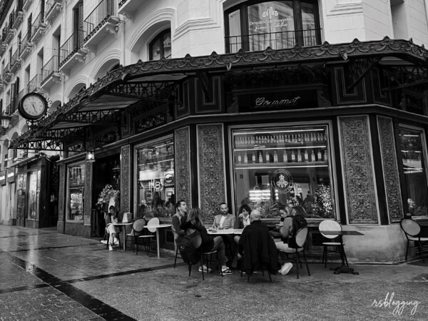 Grupo de personas sentadas en la terraza de una cafetería gourmet en una esquina señorial, bajo una marquesina con reloj y fachada decorada