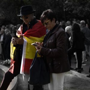 Pareja mayor consulta sus teléfonos móviles durante una concentración en la vía pública, con una bandera de España sobre los hombros de él.