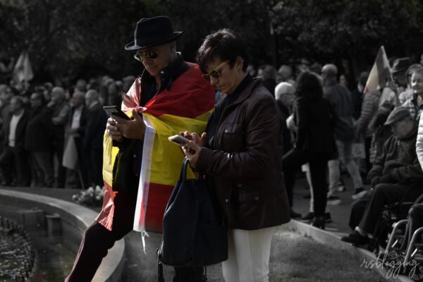 Pareja mayor consulta sus teléfonos móviles durante una concentración en la vía pública, con una bandera de España sobre los hombros de él.