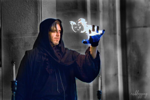 Artista vestido con túnica oscura realizando un espectáculo de magia con fuego azul en la palma de su mano.
