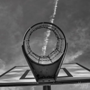 Aro de baloncesto visto desde abajo con red de cadenas, alineado con una estela en el cielo que atraviesa el centro del aro.