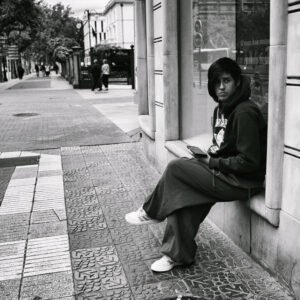 Persona joven sentada en la acera junto a un escaparate en una calle semivacía de Logroño, con expresión seria y un móvil en la mano.