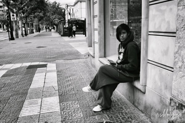 Persona joven sentada en la acera junto a un escaparate en una calle semivacía de Logroño, con expresión seria y un móvil en la mano.