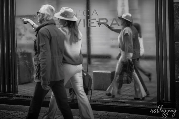 Pareja caminando por la calle, mientras la mujer señala una cristalera que refleja su imagen y la del entorno con nitidez.
