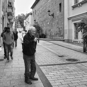 Hombre mayor caminando con bastón por una calle peatonal en blanco y negro, rodeado de transeúntes de diferentes edades.