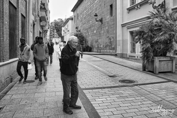 Hombre mayor caminando con bastón por una calle peatonal en blanco y negro, rodeado de transeúntes de diferentes edades.
