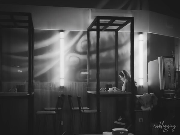 Joven sentada sola en una mesa de cafetería con auriculares puestos, concentrada en escribir o leer, bajo una tenue luz interior.